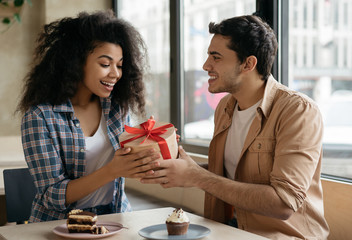 Attractive Indian man giving gift dox to beautiful African American woman for birthday. Lovely couple sitting together in cafe, romanic date. Valentines day concept