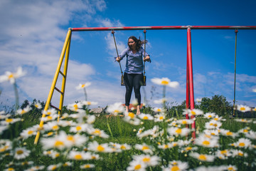 Young brunette woman is swinging on a swing, with a field of daisies (anthemidae) in the foreground on a beautiful spring sunny day. Concept of natural playfulness outdoor.