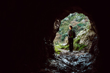 Young girl with backpack observes the landscape after going through a natural tunnel in the mountains. Concept of healthy life and adventures.