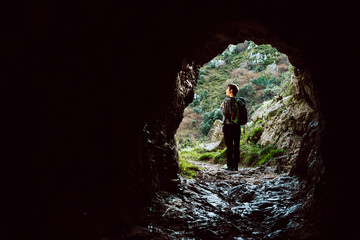Obraz premium Young girl with backpack observes the landscape after going through a natural tunnel in the mountains. Concept of healthy life and adventures.