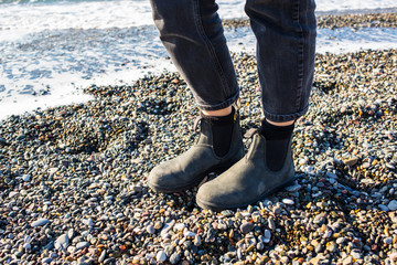 Chelsea boots classic black leather rubber sole. Focus on legs of hipster woman wearing denim black trousers. Shot on gravel beach with sea in background