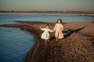 happy cute girls sisters run along the sandy seashore, summer and sunset, girls in dresses and hold hands