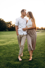 Happy couple in love in casual wear walks barefoot on the grass at sunset and hugging