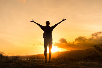Happy female standing on a mountain with arms in the air facing the sunset. 