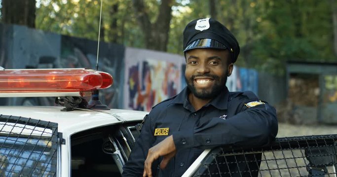 Portrait of handsome African American young policeman in uniform and cap. Front view of police officer smiling at camera outdoors. Cop on the background with police car and old lane.
