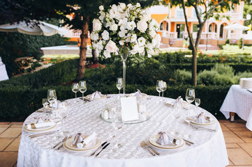Round table set for meal. White tablecloth, empty plates, forks and knives. Fresh flowers of roses, as a decoration of a wedding table.