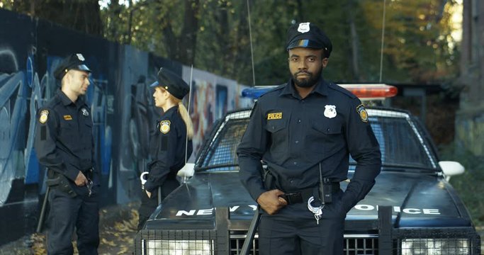 Portrait of serious African American handsome policeman in uniform looking at camera outdoor. Police officer standing and leaning on car. Caucasian colleagues talking on background.
