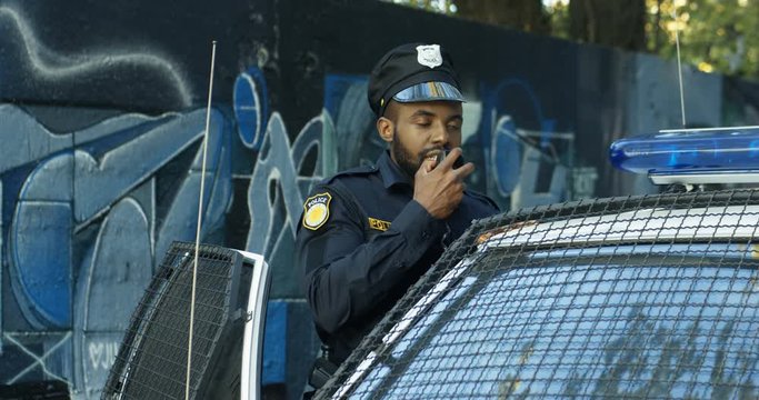 African American young cop in hat and uniform working at patrolling. Policeman standing at his police car and speaking in walkie-talkie. Cop talking in ratio.