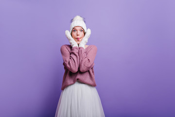 Funny surprised girl in winter attire touching her face. Studio shot of wonderful white woman in woolen clothes expressing amazement.
