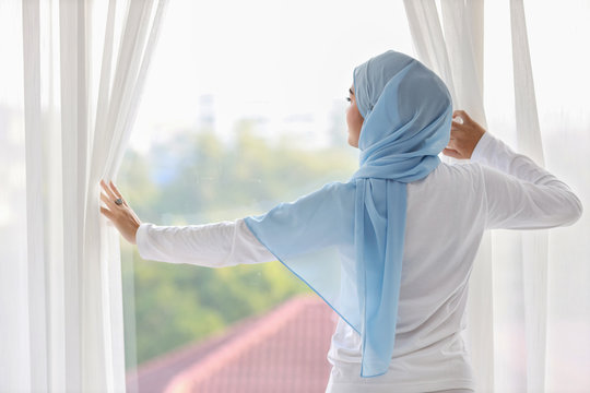 Rear view beautiful asian muslim woman wearing white sleepwear, stretching her arms after getting up in the morning at sunrise. Cute young girl with blue hijab standing and relaxing while looking away