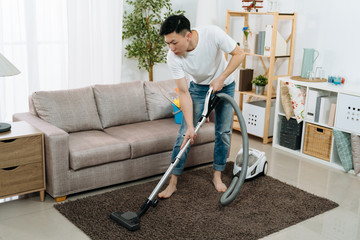Young asian japanese man cleaning carpet with vacuum cleaner at home living room. handsome guy doing housework in bright modern apartment. male household hardworking prepare tidy up for new year.