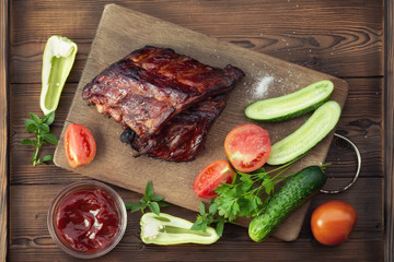 Barbecue pork ribs on a wooden cutting board with ketchup and vegetables, top view, flat lay