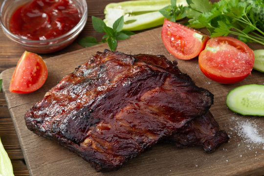 Barbecue pork ribs on a wooden cutting board with ketchup and vegetables