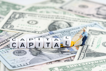 Miniature people man pulling forklift with cube small block building the word Capital on pile of US Dollar banknote money using as capital market, capitalism or global financial concept