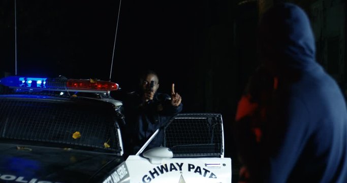 Back view of the criminal at night outdoors. African American police officer pointing pistol at offender. Cop standing at police car in dark street with gun. Rear.