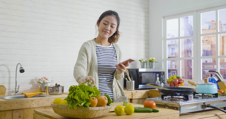 young happy asian japanese smiling woman wear sweater looking recipe in mobile phone while standing in kitchen. Vegetable salad dieting healthy lifestyle concept. girl cooking at home prepare food