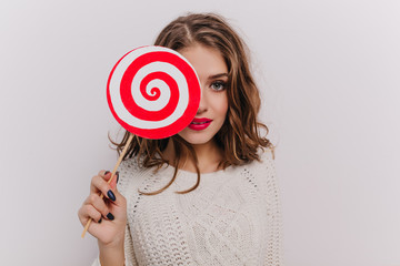 Shot of young attractive girl with wavy hair holding huge white and red lollipop on isolated background