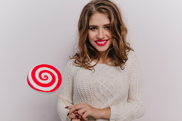 Nice girl with blue eyes and red lipstick is smiling, looking at camera and holding lollipop in her hands on white background