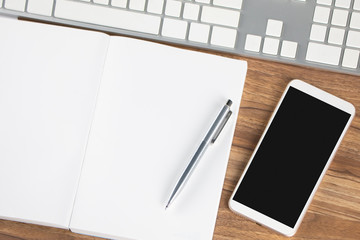 Modern office desk with blank screen smartphone, notebook and computer keyboard on wooden background with copy space. Top view. Education, work space and business concept.