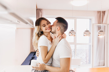 Brunette man holding cup of coffee and kissing wife. Brown-haired guy in white t-shirt enjoying weekend morning with charming european girl.