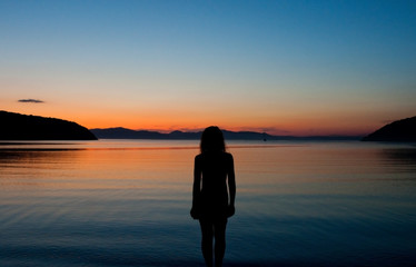Fototapeta premium black silhouette of a woman on the background of a stunning orange and blue sunset on the sea, the sunset sky is reflected in the water