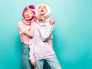 Two young sexy hipster girls in  wigs and red lips.Beautiful trendy women in summer clothes.Carefree models posing near blue wall in studio.Positive female going crazy.Gives air kiss