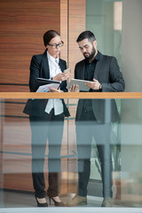 Busy young colleagues in formalwear standing in office corridor and synchronizing devices while...