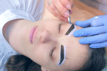 Obraz na płótnie Canvas Professional beautician plucking eyebrows with tweezers to woman in beauty salon during tint eyebrow procedure, face closeup. Girl lying with closed eyes and brown natural henna on brows.