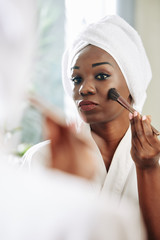 Vertical portrait of beautiful Black woman standing in front of mirror applying blusher on her face using brush