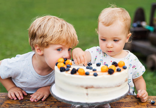 Two Toddler Children With Birthday Cake Outdoors In Garden In Summer.