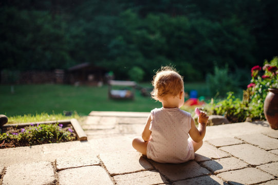 A rear view of toddler girl sitting outdoors on patio in summer.