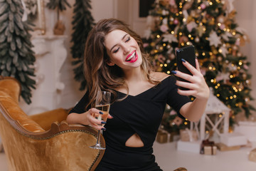 Charming woman with trendy manicure making selfie with champagne near christmas tree. Indoor portrait of cute white female model in party attire taking picture of herself in holiday.