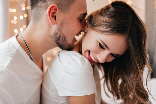 White young woman laughing while husband kissing her neck. Indoor shot of black-haired bearded guy playfully posing with girlfriend.