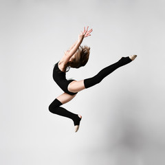 Naklejka premium Young girl gymnast in black sport body and uppers jumping and making dymnastic pose in air over white background