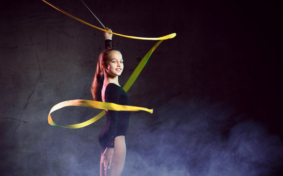 portrait of beautiful young brunette woman gymnast training calisthenics exercise with yellow ribbon on dark smoke studio background.