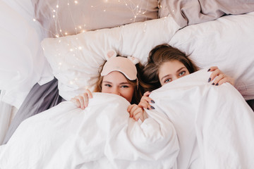 Good-looking caucasian girls hiding under white blanket. Indoor portrait of amazing young sisters posing in morning.