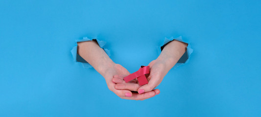 Red ribbon is symbol of fight against AIDS or disease cancer of female breast. Girl's hand holds a red ribbon on a blue torn papper background. Copy space aside for your advertising content