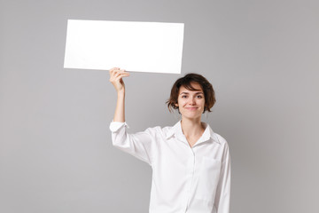 Smiling pretty young business woman in white shirt posing isolated on grey background. Achievement career wealth business concept. Mock up copy space. Hold white blank sign board with place for text.