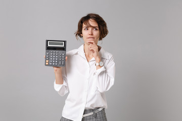 Pensive young business woman in white shirt posing isolated on grey wall background in studio. Achievement career wealth business concept. Mock up copy space. Hold calculator put hand prop up on chin.