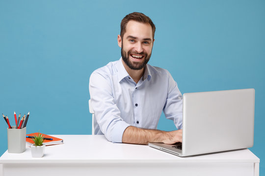 Smiling young bearded man in light shirt sit at white desk isolated on pastel blue wall background. Achievement business career concept. Mock up copy space. Working on project with laptop pc computer.