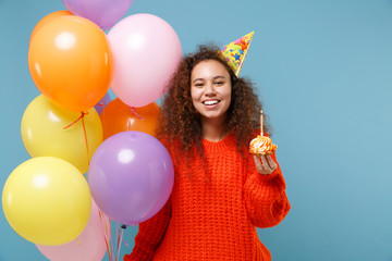 Smiling young african american girl in casual orange knitted clothes, birthday hat isolated on pastel blue background. Holiday party concept. Celebrating, hold colorful air balloons, cake with candle.