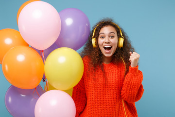 Fototapeta na wymiar African american girl in orange knitted clothes isolated on blue background. Birthday holiday party concept. Celebrating hold colorful air balloons listen music with headphones doing winner gesture.