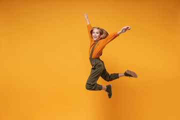 Funny little kid girl 12-13 years old in turtleneck, jumpsuit isolated on orange yellow wall background children studio portrait. Childhood lifestyle concept. Mock up copy space. Jumping rising hands.