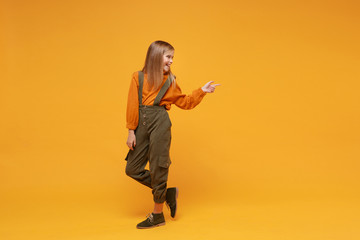 Side view of smiling little blonde kid girl 12-13 years old in turtleneck, jumpsuit isolated on orange yellow background. Childhood lifestyle concept. Mock up copy space. Pointing index finger aside.