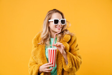Smiling young blonde woman girl in yellow fur coat, dark sunglasses posing isolated on orange background in studio. People lifestyle concept. Mock up copy space. Holding plastic cup of cola or soda.