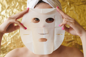 Young woman apply cosmetic fabric facial mask on golden background. Face peeling mask with charcoal, spa beauty treatment, skincare, cosmetology. Close up