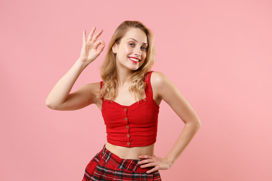 Smiling young blonde woman girl in red sexy clothes posing isolated on pastel pink wall background studio portrait. People sincere emotions lifestyle concept. Mock up copy space. Showing OK gesture.