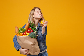 Smiling woman in denim clothes isolated on yellow orange background. Delivery service from shop or restaurant concept. Hold brown craft paper bag for takeaway mock up with food products, bell pepper.