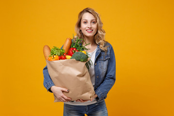 Smiling young woman in denim clothes posing isolated on yellow orange background. Delivery service from shop or restaurant concept. Hold brown craft paper bag for takeaway mock up with food products.
