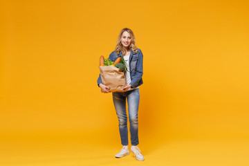 Smiling young woman in denim clothes isolated on yellow orange wall background. Delivery service from shop or restaurant concept. Hold brown craft paper bag for takeaway mock up with food products. © ViDi Studio